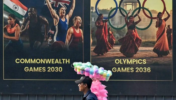 Vendedor de algodão doce passa por cartazes dos Jogos da Commonwealth e dos Jogos Olímpicos, em Ahmedabad, na Índia 
             -       
        Shammi Mehra - 26.nov.2025/AFP