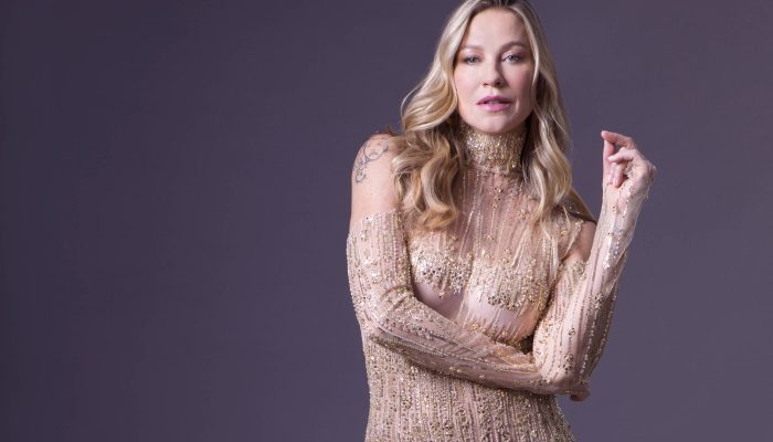 Mulher loira em pé com macacão justo bege coberto por lantejoulas brilhantes. Ela usa botas combinando e está contra fundo cinza escuro.